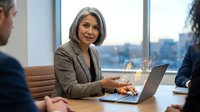 Mature businesswoman with gray hair working on laptop during corporate meeting in modern office conference room with city view.