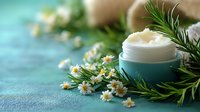 Natural organic cream in turquoise jar with fresh chamomile flowers and rosemary sprigs on textured blue background, wellness and skincare concept.