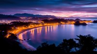 Aerial view of illuminated coastal city at twilight with glowing lights reflecting on curved bay waters against dramatic purple and orange sunset sky.