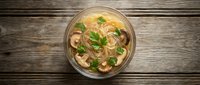 Asian noodle soup with mushrooms and herbs in glass bowl on rustic wooden background, perfect for restaurant menus and food blogs.