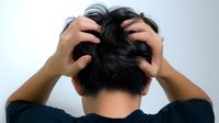 Back view of Asian man scratching head with both hands against light background, showing dark short hair and black shirt, expressing frustration or confusion.