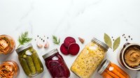 Assorted preserved foods in glass jars including pickled vegetables, fermented cabbage, beets and spreads arranged on white marble background.