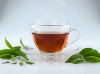 Clear glass teacup with amber tea on saucer surrounded by fresh green tea leaves on white background for beverage and wellness designs.