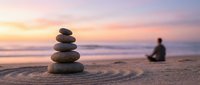 Zen stone stack on beach sand with blurred person meditating at sunset, peaceful mindfulness and balance concept for wellness and spiritual practice.