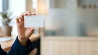 Close-up of male hand holding blank white business card in modern office environment with blurred background, suitable for corporate branding and professional identity concepts.