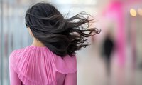 Young Asian woman with flowing black hair in pink pleated blouse walking away, motion blur effect captures wind-blown hair against soft bokeh background.