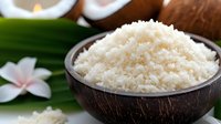 Fresh grated coconut in natural coconut shell bowl on green palm leaf with tropical flower and whole coconuts in soft focus background.