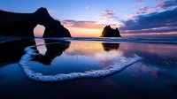 Dramatic coastal sunset with natural rock arch and sea stacks reflected in wet sand. Colorful sky with pink and orange clouds over ocean waves at twilight.