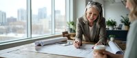 Mature businesswoman architect reviewing blueprints at office desk with city view. Professional consultation meeting for construction project planning.