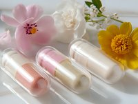 Transparent capsules filled with shimmering pearl powder and pink pigments alongside delicate pink, white, and yellow flowers on light background.