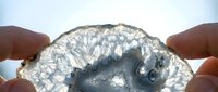 Hands holding translucent gray agate slice with intricate crystalline patterns and natural formations against bright sky background for jewelry design.