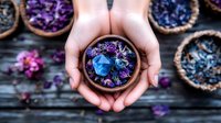 Hands holding wooden bowl with vibrant dried flowers and blue petals against rustic dark background, surrounded by scattered purple and blue botanical elements.