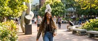 Young African American woman with dreadlocks walking confidently through urban plaza with modern sculptures and lush greenery on sunny day.