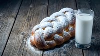 Fresh braided challah bread dusted with powdered sugar served with glass of milk on rustic wooden table for breakfast or traditional meal.