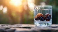 Glass of water with dried dates on wooden surface against blurred natural bokeh background with warm sunset light and copy space for Ramadan concept.