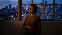 Young woman in cozy sweater standing by large windows overlooking illuminated city skyline at dusk, urban apartment living with warm interior lighting.