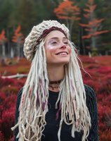 Young Caucasian woman with white dreadlocks and face glitter wearing knit beanie smiles outdoors against autumn red foliage background. Natural lifestyle portrait.
