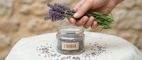 Unrecognizable person holding fresh lavender bunch above glass jar filled with dried lavender buds on stone surface for aromatherapy and herbal wellness products.