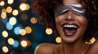 Woman with curly hair wearing silk blindfold laughs joyfully against blurred bokeh lights, expressing happiness and excitement for celebration or party themes.