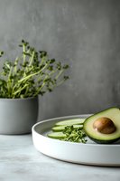 Fresh avocado half with cucumber slices and microgreens on white ceramic plate against concrete background, styled for healthy food photography.