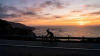 Cyclist riding along coastal road at sunset with dramatic orange and purple sky, ocean and cliffs in background, suitable for sports and travel content.