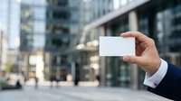 Businessman in dark suit holding blank white business card outdoors in urban downtown setting with modern buildings, professional networking and corporate identity concept.