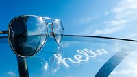 Aviator sunglasses resting on a reflective surface with hello text, blue sky and clouds in background, summer vibes.