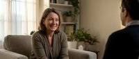 Middle-aged Caucasian woman smiling during consultation meeting with professional advisor in modern living room setting for therapy or business discussion.