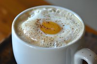 Creamy latte coffee topped with silky milk foam and sprinkled cocoa powder in white ceramic cup, shallow depth of field creates soft background blur.