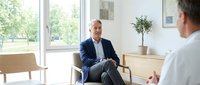 Middle-aged Caucasian male professional in navy blazer and light shirt sitting in modern office chair during business meeting or consultation with colleague, bright natural light from windows, contemporary workplace interior.