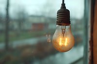 Glowing vintage light bulb hanging against blurred rainy window background, creating moody atmospheric ambiance with warm illumination and bokeh effect.