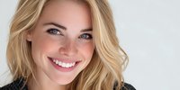 Young Caucasian woman with blonde hair, blue eyes and freckles smiling at camera against light gray background, conveying happiness and confidence.