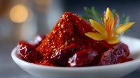 Red chili paste garnished with yellow lily flower in white ceramic bowl, surrounded by whole red chilies, creating vibrant contrast against dark background.