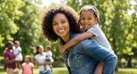 African American mother and daughter enjoying piggyback ride in sunny park with family gathering in background during summer outdoor recreation.