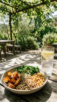 Healthy vegan bowl with quinoa, chickpeas, roasted sweet potato and kale served outdoors with lemon water on a sunny wooden table.
