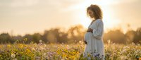 Pregnant African American woman in white dress standing in wildflower field during golden hour sunset, maternity photography concept for expecting mothers.