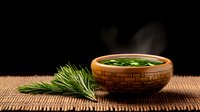 Fresh rosemary sprigs beside wooden bowl with aromatic herbal tea on bamboo mat against dark background, creating zen atmosphere for wellness and relaxation.