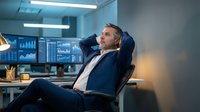 Relaxed businessman in suit leaning back in office chair with hands behind head, multiple monitors displaying financial data charts in modern workplace.