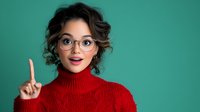 Young Asian woman with glasses and curly hair wearing red turtleneck sweater showing eureka gesture with raised finger against turquoise background, expressing bright idea moment.