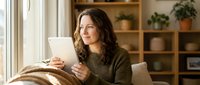 Woman with long dark hair and medium complexion, wearing olive green ribbed sweater, sitting by window holding tablet device, smiling while working or browsing. Cozy home office setting with wooden shelving and potted plants.