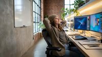 Young Caucasian woman relaxing at creative workspace with dual monitors, hands behind head, enjoying a break in modern loft office with brick walls.