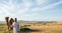 Man in white traditional clothing standing with camel in desert landscape with mountains and blue sky, suitable for travel and cultural content.
