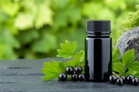 Black supplement bottle with fresh blackcurrants and green leaves on dark stone surface against blurred garden background, natural wellness concept.