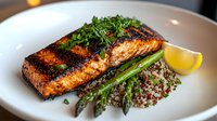 Grilled salmon fillet with charred crust, served on quinoa pilaf, garnished with fresh herbs and asparagus spears, accompanied by lemon wedge on white plate.