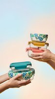 Hands holding colorful glass food storage containers with botanical lids against pastel gradient background for sustainable kitchen organization.