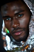 Close-up portrait of young African American male model with striking blue eyes wrapped in reflective metallic material against dark background with bokeh lights.