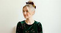 Blonde woman with upswept hairstyle wearing gold hair clip and dark green velvet floral top, looking to side against neutral background for fashion and beauty content.