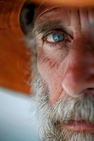 Close-up portrait of senior man with bright blue eyes and gray beard wearing orange cap, showing wisdom and life experience in dramatic lighting.
