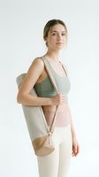 Young woman with blonde hair and fair complexion wearing sage green sports bra and cream leggings, carrying beige yoga mat bag over shoulder against white background for fitness and wellness content.