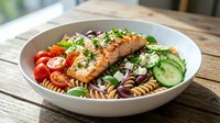 Grilled salmon fillet with pasta salad, cherry tomatoes, cucumber and fresh herbs in white bowl on wooden table for healthy Mediterranean cuisine.